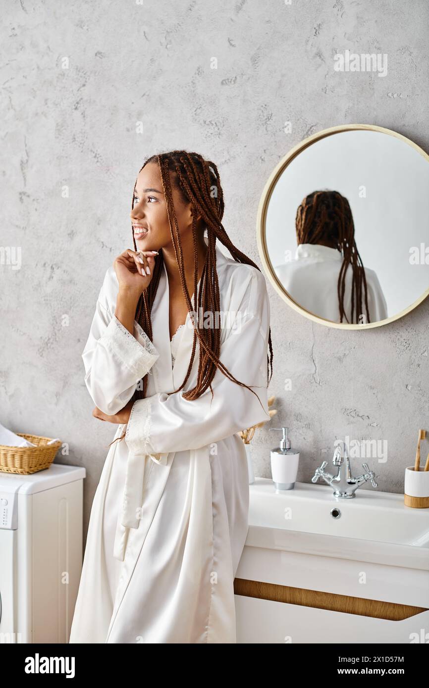 Afroamerikanerin mit Afro-Flechten steht im Bademantel vor dem modernen Badezimmerspiegel. Stockfoto