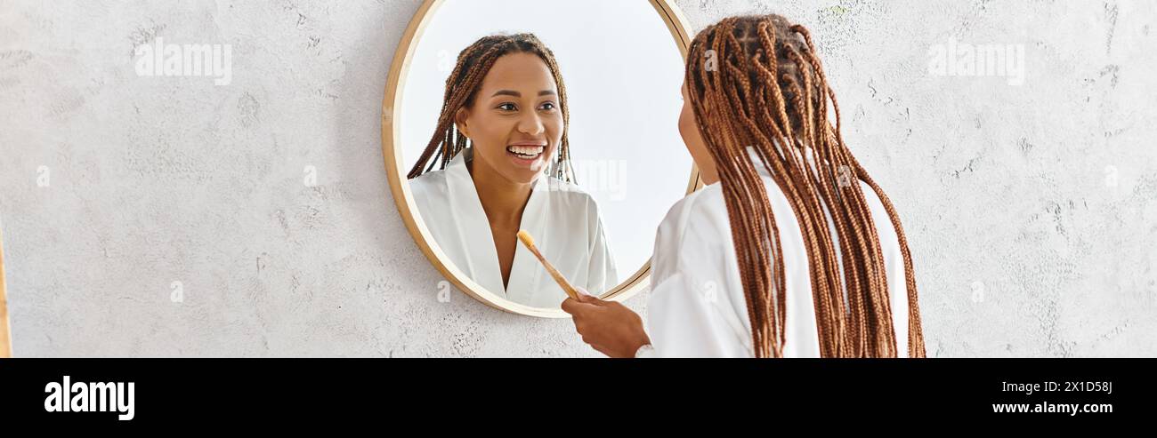Eine Frau mit Afro-Zöpfen schaut auf ihr Spiegelbild in einem Badezimmerspiegel und konzentriert sich dabei auf Selbstverständnis und Schönheit. Stockfoto
