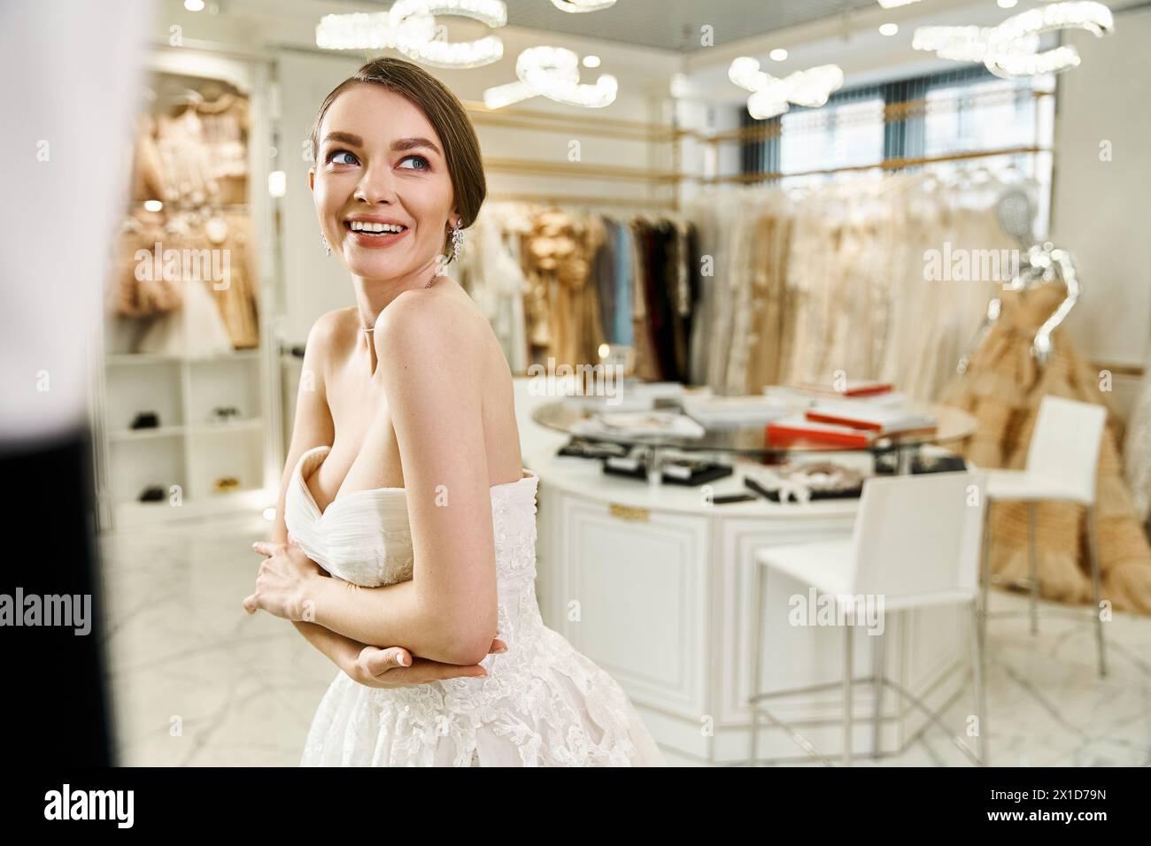 Eine junge, brünette und schöne Braut steht selbstbewusst vor einem Spiegel in einem Kleiderladen und probiert verschiedene weiße Kleider für ihren besonderen Tag an. Stockfoto