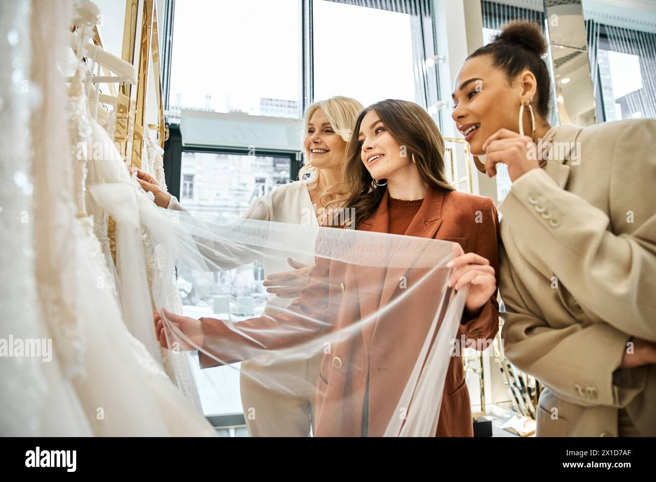 Eine junge Braut, ihre Mutter und ihr bester Freund stehen vereint vor einer Auswahl von Kleidern. Stockfoto