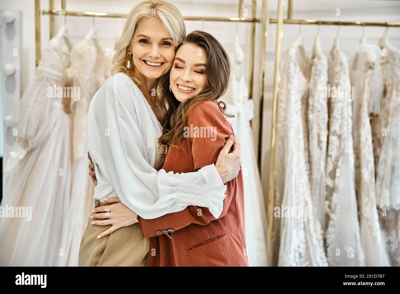 Zwei Frauen umarmen sich vor Brautkleidern, eine junge Braut und ihre Mutter teilen einen Moment der Freude und Liebe. Stockfoto