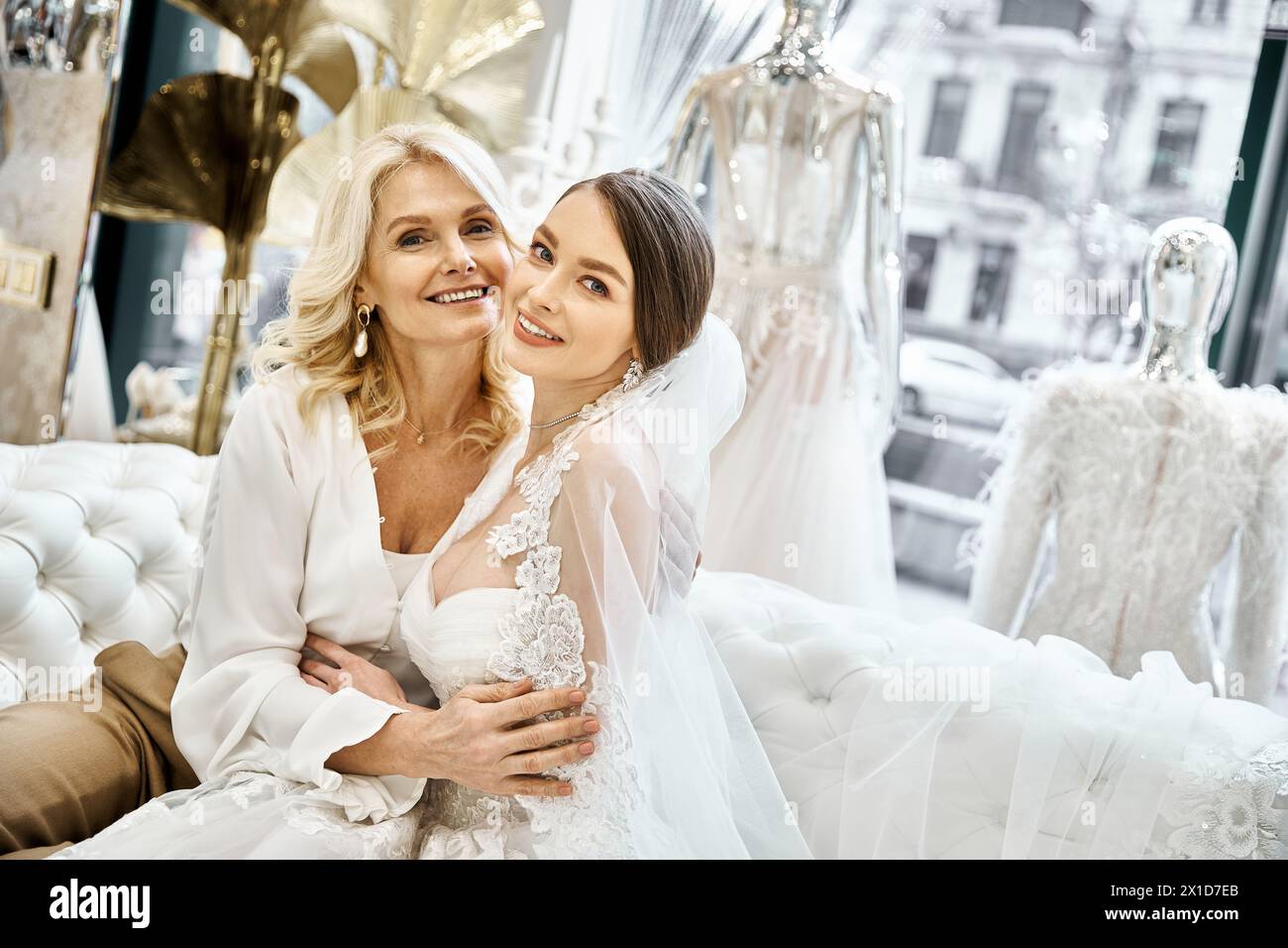 Eine junge brünette Braut und ihre blonde Mutter mittleren Alters sitzen zusammen in Brautkleidern in einem Brautsalon. Stockfoto