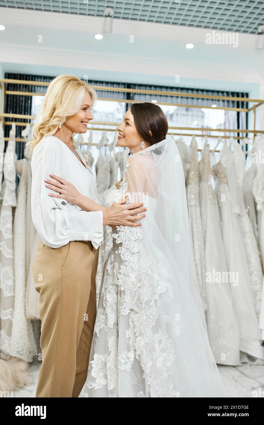 Eine junge brünette Braut in einem Hochzeitskleid und ihre Mutter mittleren Alters stehen Seite an Seite in einem Brautsalon. Stockfoto