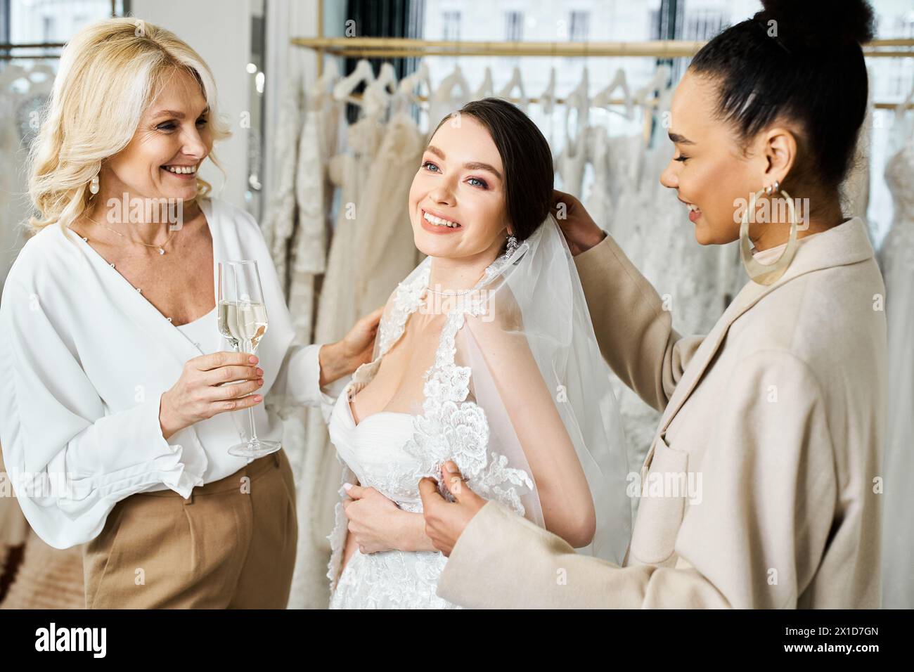Eine junge Braut, ihre Mutter und die Brautjungfer stehen anmutig in verschiedenen Kleidern in einem Brautsalon. Stockfoto