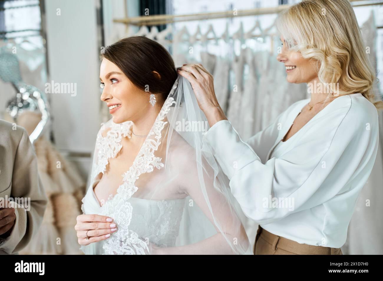 Die junge Braut in einem weißen Kleid und die andere ihre Mutter stehen neben einem Kleiderständer in einem Brautsalon. Stockfoto