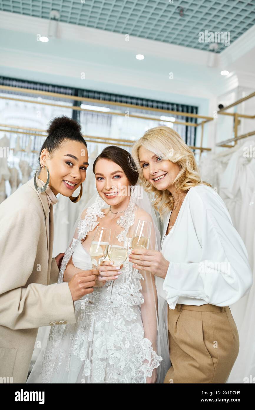 Eine junge Braut im Hochzeitskleid steht zwischen ihrer Mutter mittleren Alters und ihrer besten Freundin in einem Brautsalon und lächelt warm. Stockfoto