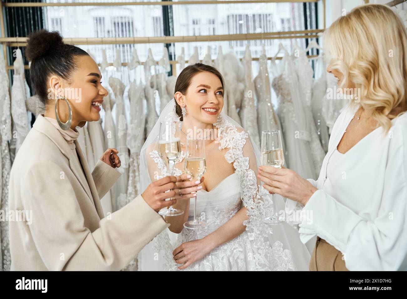 Zwei Bräute in Hochzeitskleidung und eine Frau mit Sektflöten vor einem Kleiderständer im Brautsalon. Stockfoto