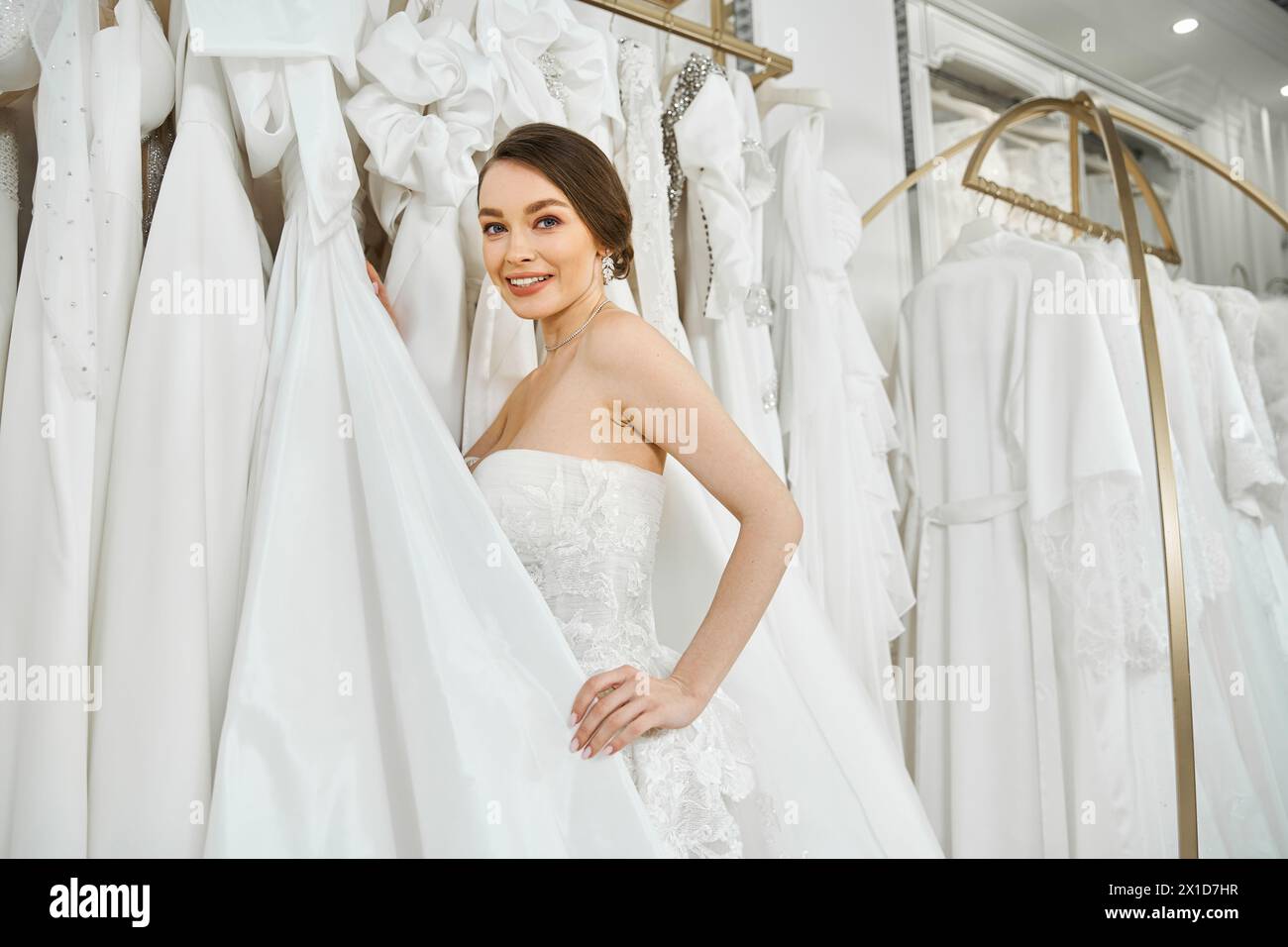 Eine junge brünette Braut steht vor einem Regal mit weißen Kleidern und wählt sorgfältig ihr perfektes Hochzeitskleid aus. Stockfoto