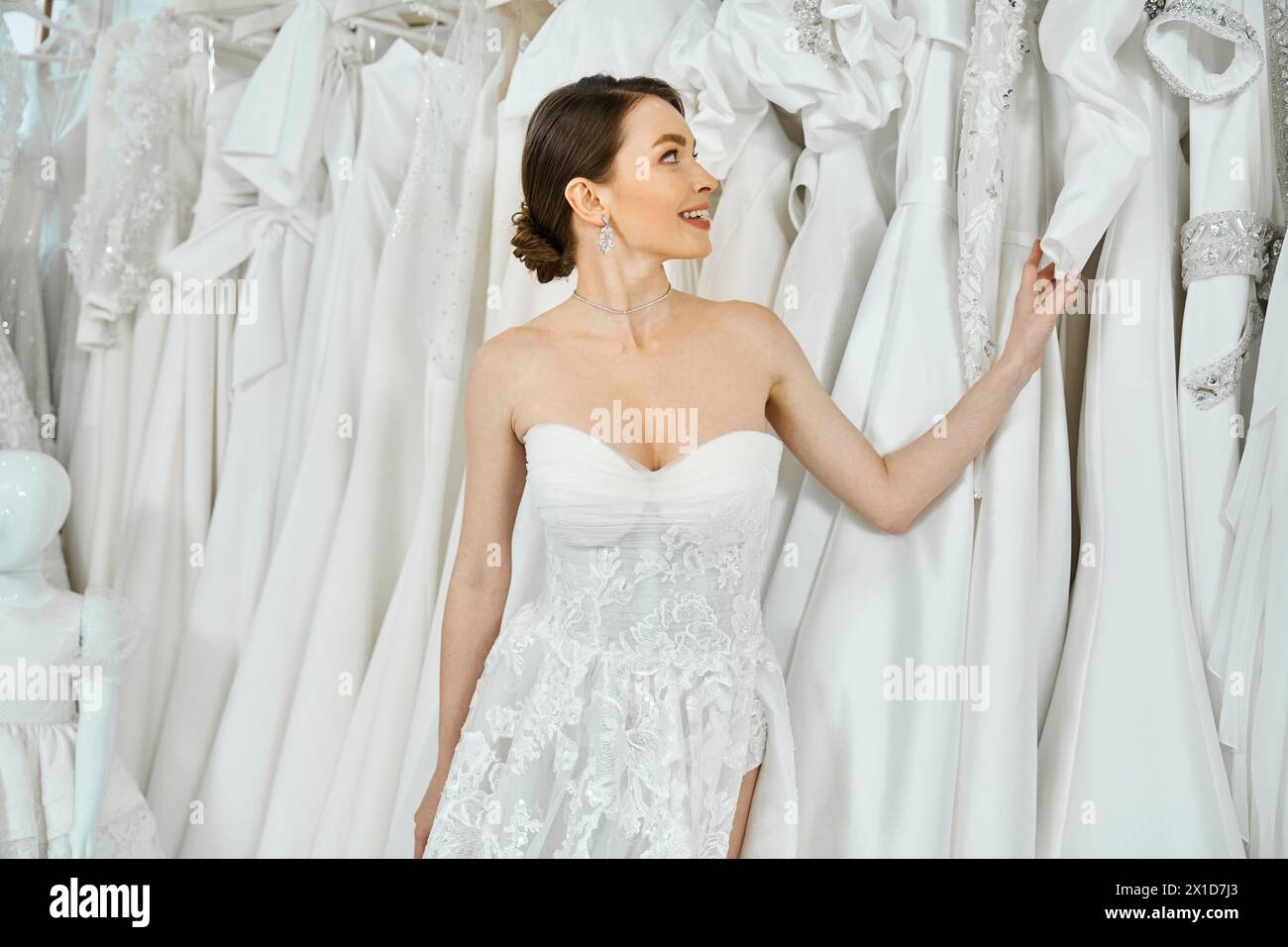 Eine junge, brünette Braut, die in einem Hochzeitssalon zwischen verschiedenen Kleidern steht. Stockfoto