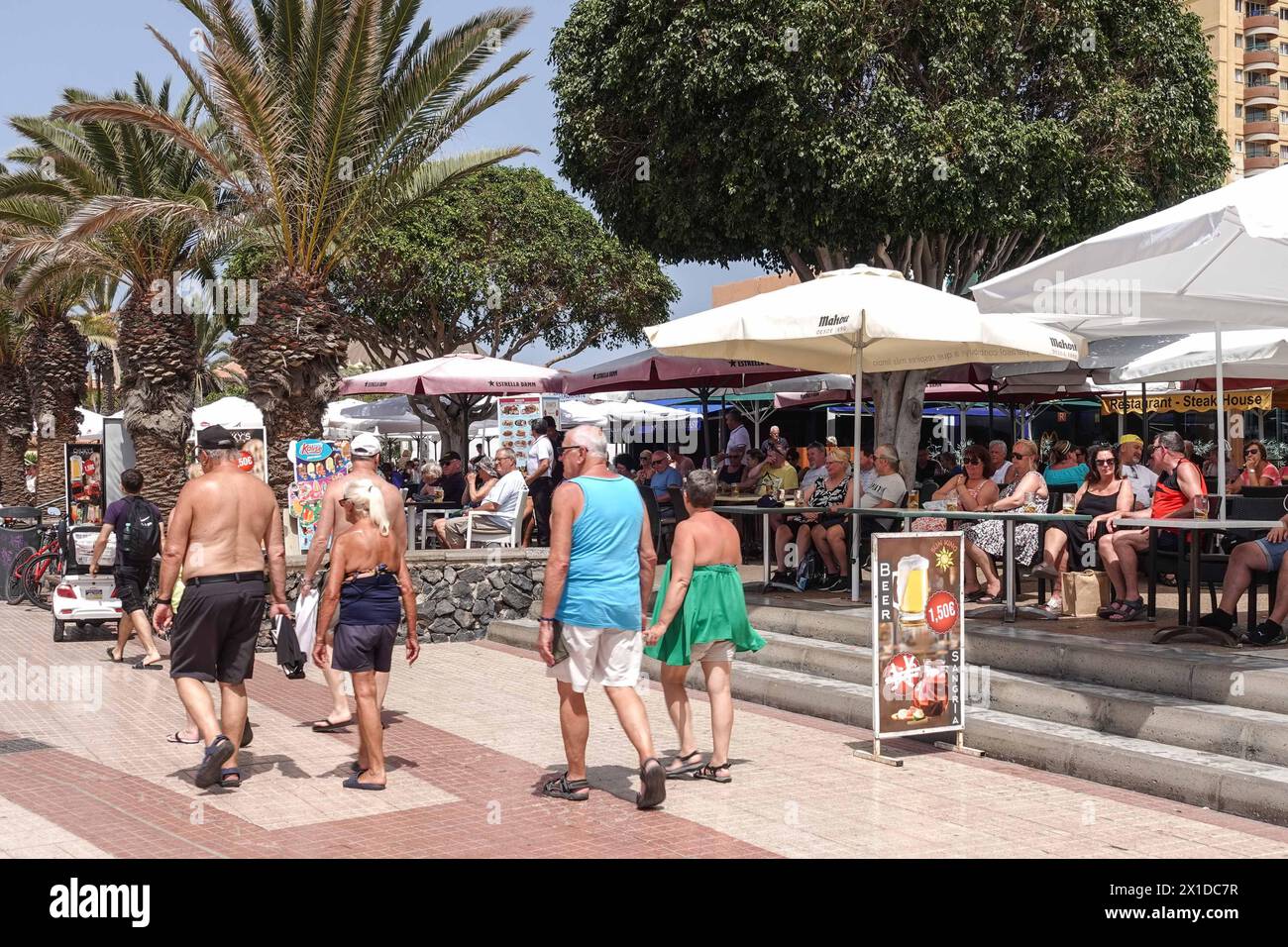 Los Cristianos, Teneriffa, 16. April 2024 - Touristen trinken in englisch freundlichen Strandbars, wo ein Bier 1,50 Euro kostet. - Touristen laufen an Anti-Touristen-Plakaten entlang des Strandes in Los Cristianos vorbei. Auf dem Poster steht, dass die Kanarischen Inseln eine Grenze haben. Die gelben Schilder werben für den anti-touristischen Protest am Samstag, den 20. April. In Santa Cruz de Teneriffa. Parken ist auch ein großes Problem in der Gegend, da unzählige Besucher Tausende von Plätzen füllen, was für die Bewohner und Besucher gleichermaßen Kopfschmerzen bereitet. Quelle: Stop Press Media/Alamy Live News Stockfoto