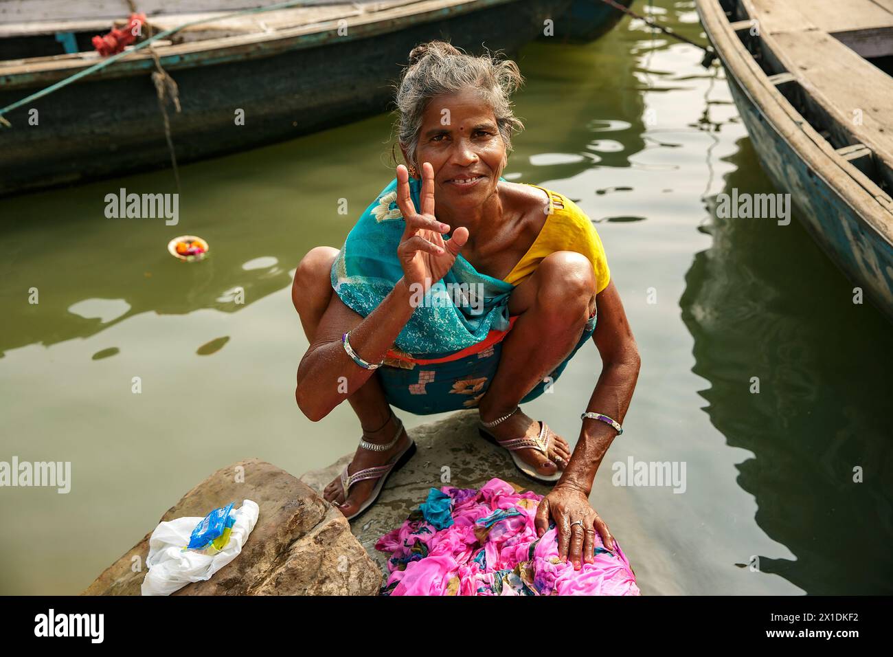 Die Frau hebt das Friedenszeichen auf, während sie im Ganges bei Varanasi, Indien, ein Kleidungsstück wäscht. Stockfoto