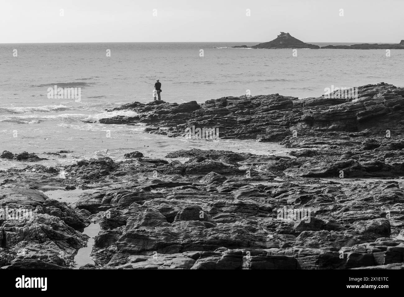 Ein Schwarzweißfoto eines einsamen Fischers, der auf den Felsen fischt, Kenneggy Sand oder Kenneggy Sands, einem abgelegenen Strand im Südwesten von Cornwall, England, Großbritannien Stockfoto