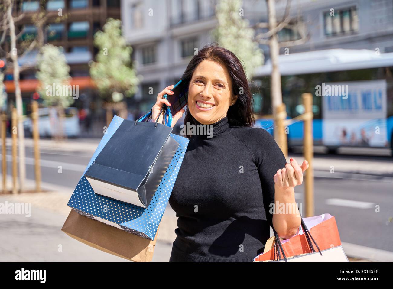 Eine elegante Frau mittleren Alters, die mit dem Smartphone telefoniert und ein Taxi mit Einkaufstaschen bestellt Stockfoto