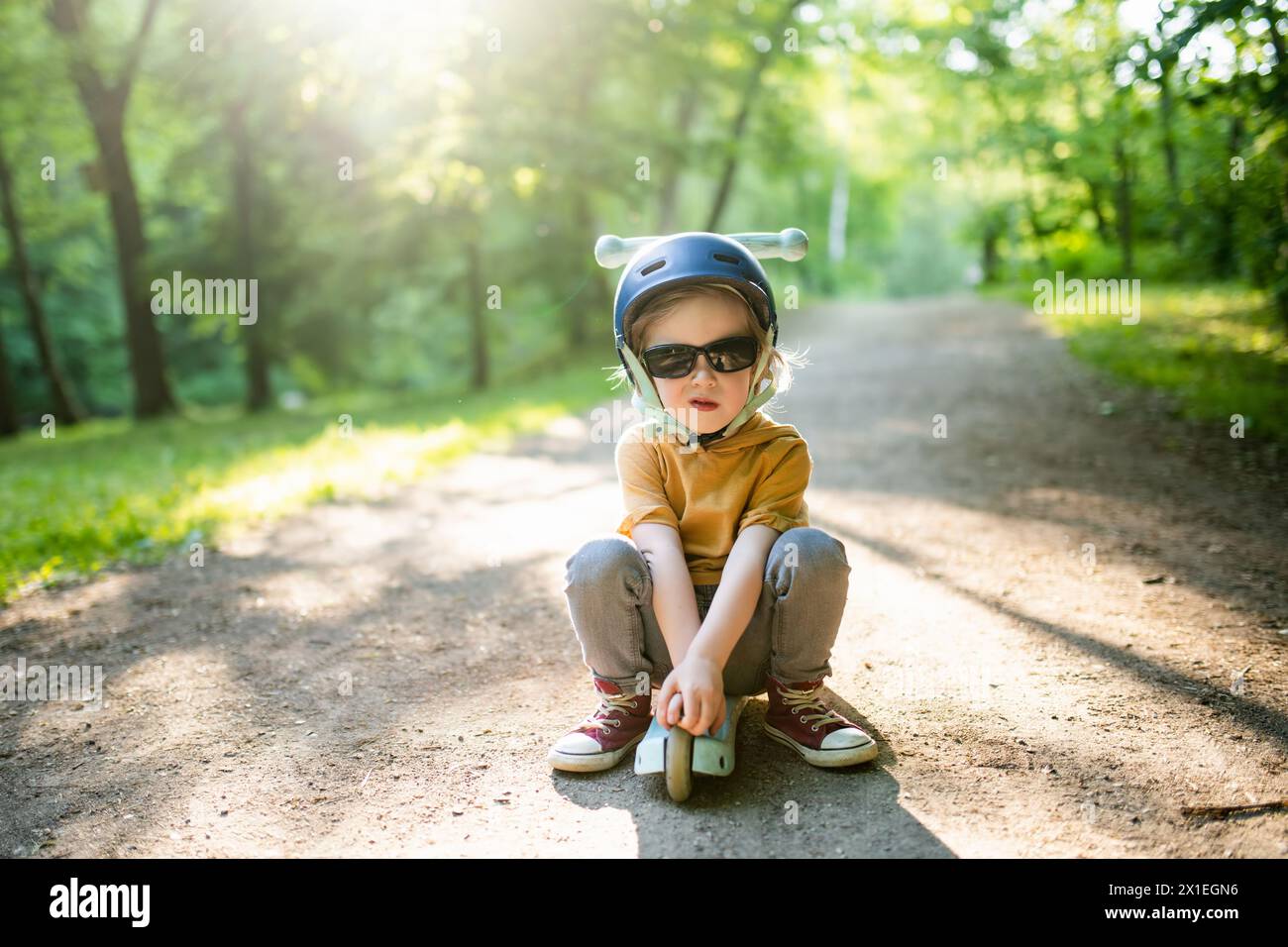 Entzückender kleiner Junge, der seinen Roller in einem Stadtpark am sonnigen Sommerabend fährt. Kleines Kind, das mit Helm auf einer Rollrolle fährt. Aktive Freizeit und Outdoo Stockfoto