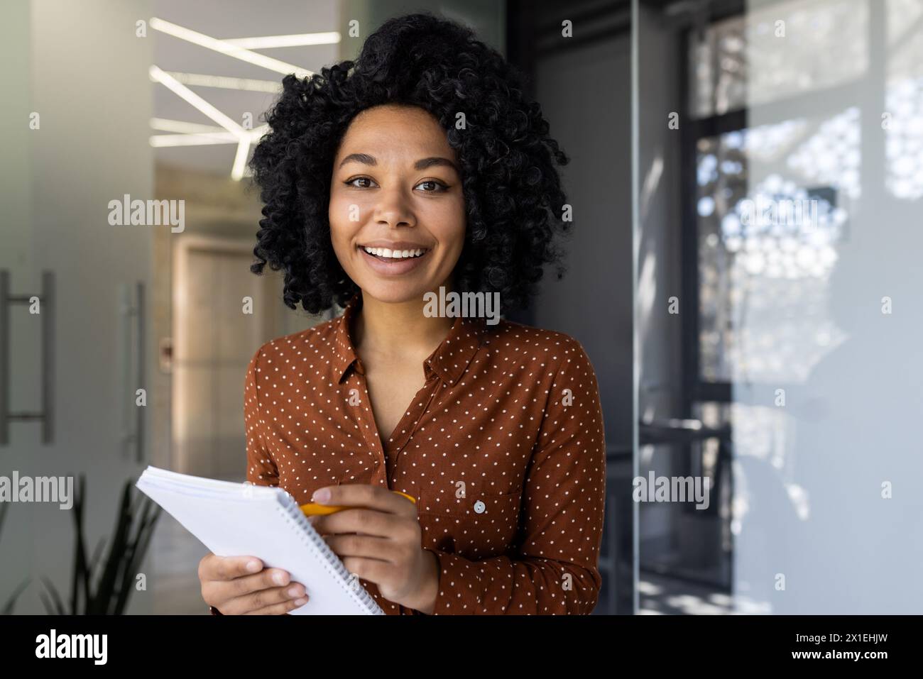 Eine fröhliche junge Frau mit lockigen Haaren nimmt an einem Online-Training Teil, macht Notizen und lächelt in einem modernen Büroumfeld. Stockfoto