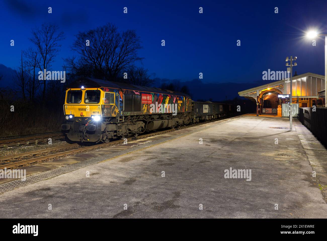 GB Rail Freight Lokomotive der Baureihe 66 66718 unter dem Vordach der Midland Railway am Bahnhof Hellifield mit einem Zug, der nachts Zuschlagstoffe transportiert Stockfoto