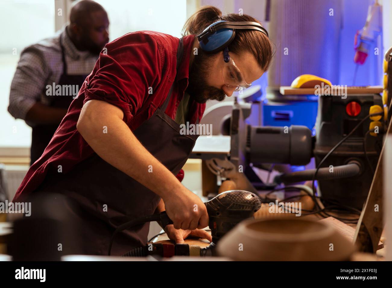 Handwerker in Tischlerei mit Sicherheits-Ohrenschützern mit Schwingschleifer auf Holz. Der Mann in der Tischlerei verwendet einen Winkelschleifer, um Holzgegenstände zu kreieren, die von einem Kollegen mit verschwommenem Hintergrund unterstützt werden Stockfoto