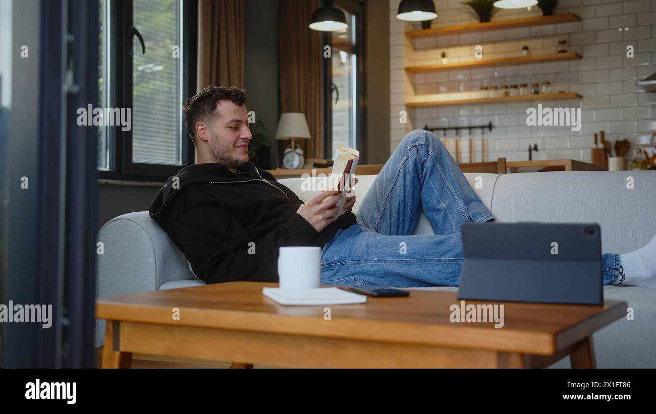 Sorgloser junger Mann verbringt das Wochenende zu Hause entspannt auf dem Sofa mit Buch lesen interessante Geschichte Genießen Sie Lieblingsliteratur. Zeit zur freien Verfügung, pas Stockfoto