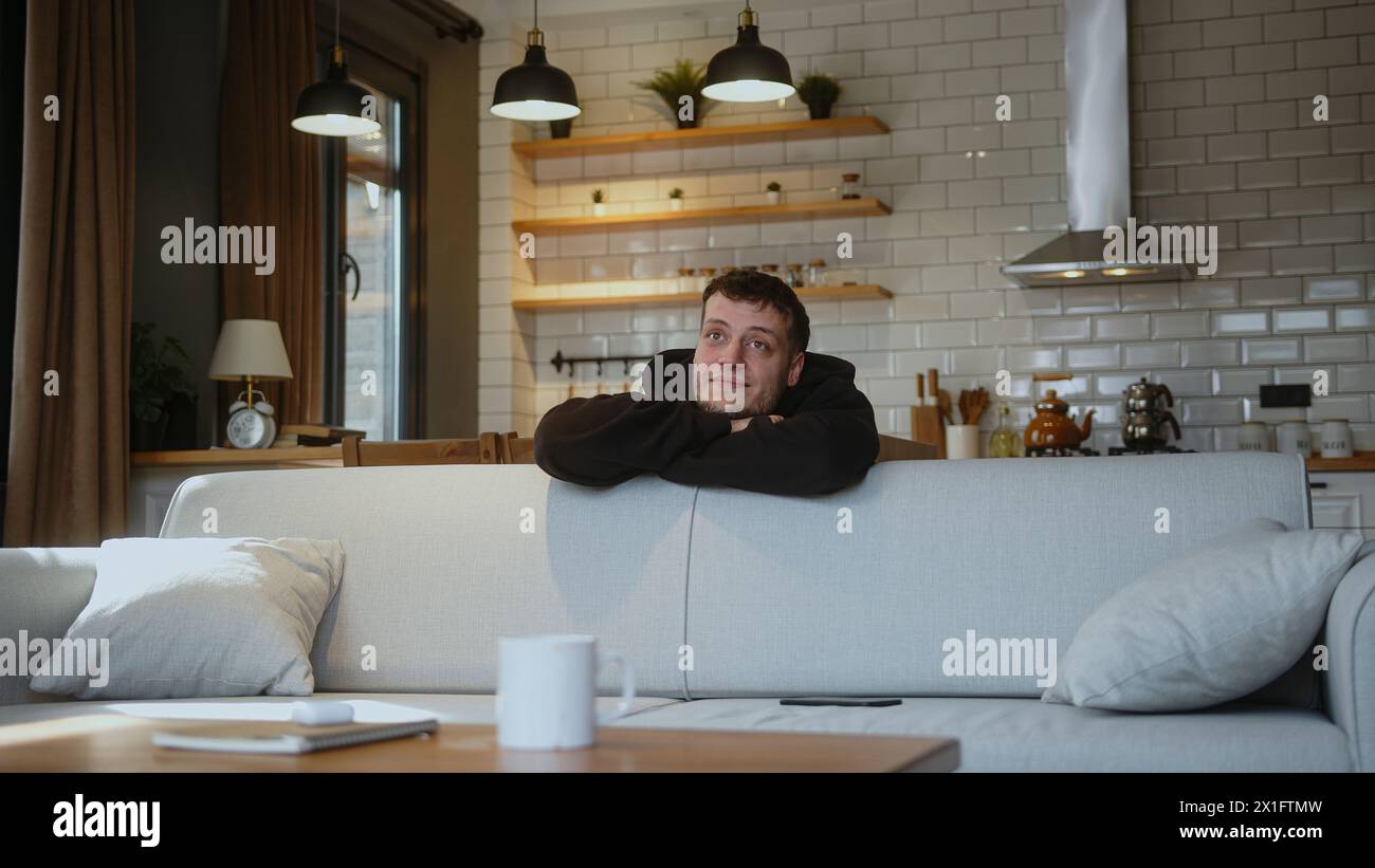 Ein lächelnder Mann lehnt sich über die Rückseite des Sofas, legt das Smartphone auf das Sofa und denkt tief in Gedanken, die zu Hause träumen. Stockfoto