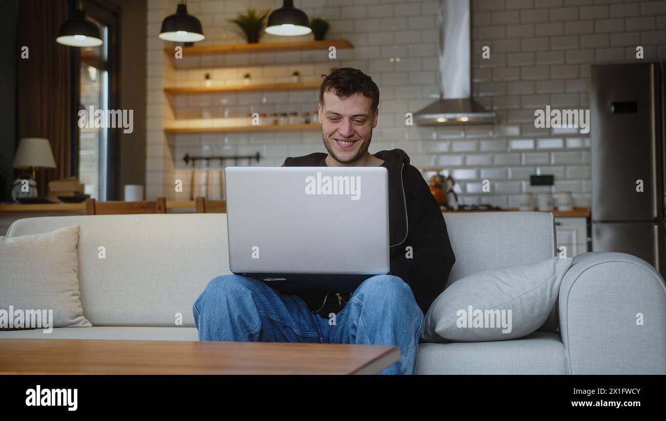 Lächelnder junger Mann, der am Laptop arbeitet, während er auf einem Sofa im Wohnzimmer sitzt. Im Internet surfen, Kaffee aus einer Tasse trinken, zu Hause Spaß haben Stockfoto