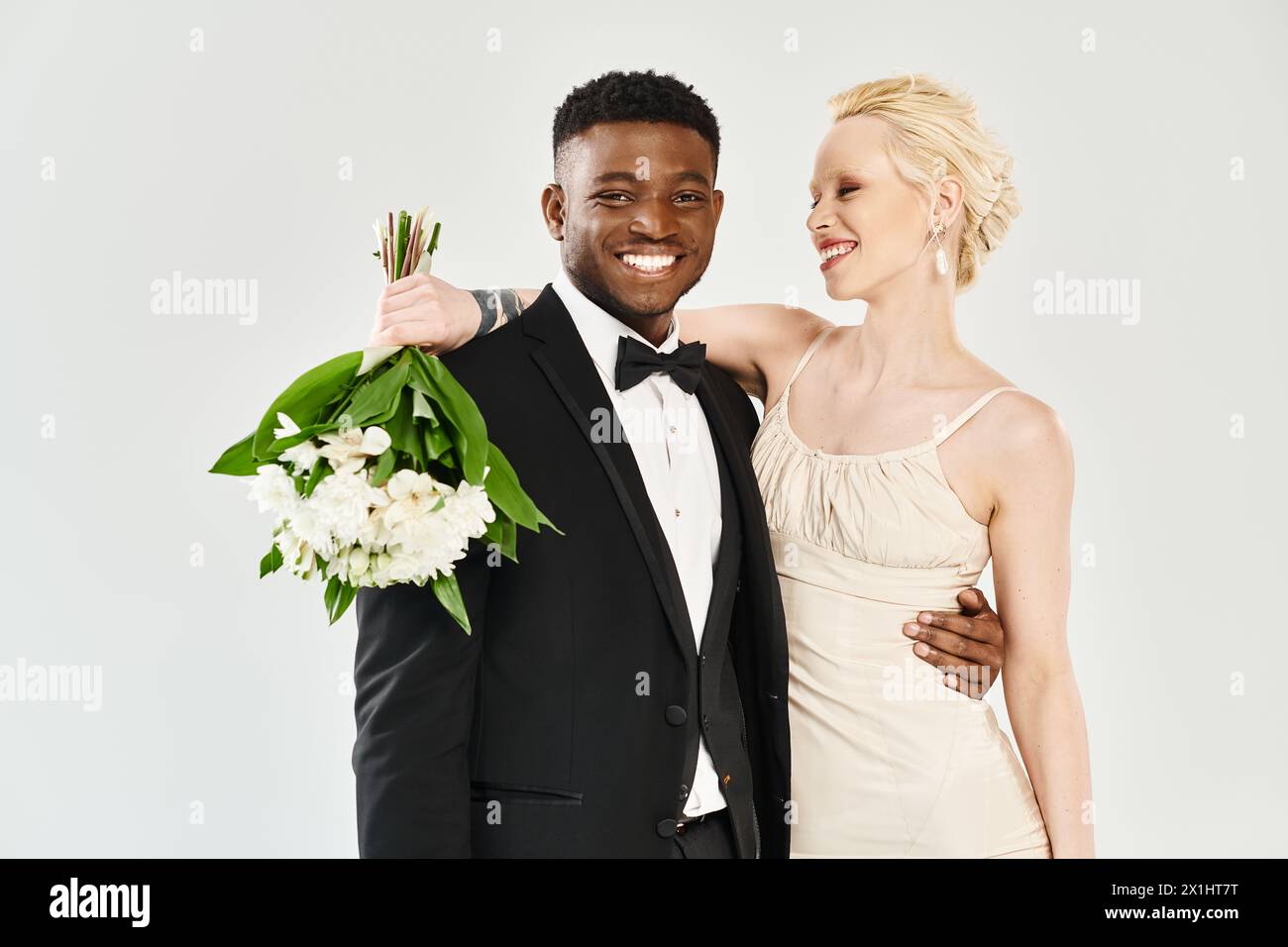 Eine wunderschöne blonde Braut in einem Hochzeitskleid und ein Afroamerikaner im Smoking stehen selbstbewusst zusammen in einem Studio. Stockfoto