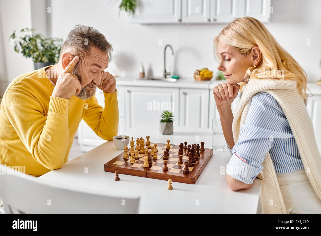 Ein reifes, liebevolles Paar in kuscheligen Hausbekleidung, das sich in einem strategischen Schachkampf an einem Tisch in ihrer Küche zu Hause engagiert. Stockfoto