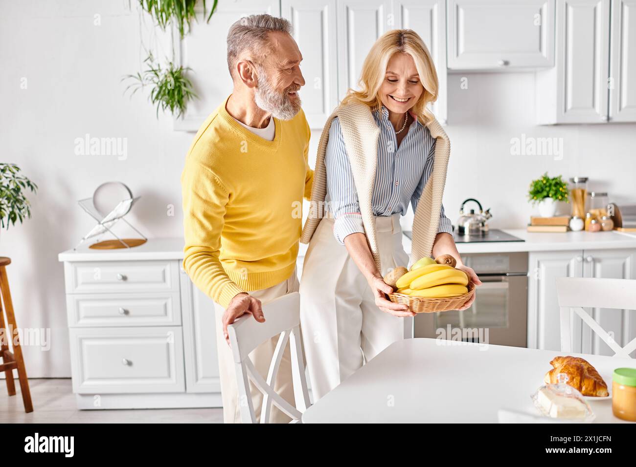 Ein reifer Mann und eine reife Frau stehen in ihrer gemütlichen Küche, halten Bananen und teilen einen süßen Moment miteinander. Stockfoto