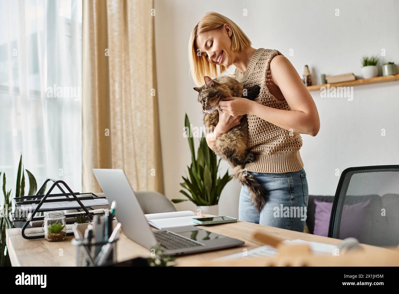 Eine stilvolle Frau mit kurzen Haaren, die ihre Katze vor einem Laptop-Bildschirm hält, vertieft in die digitale Interaktion. Stockfoto