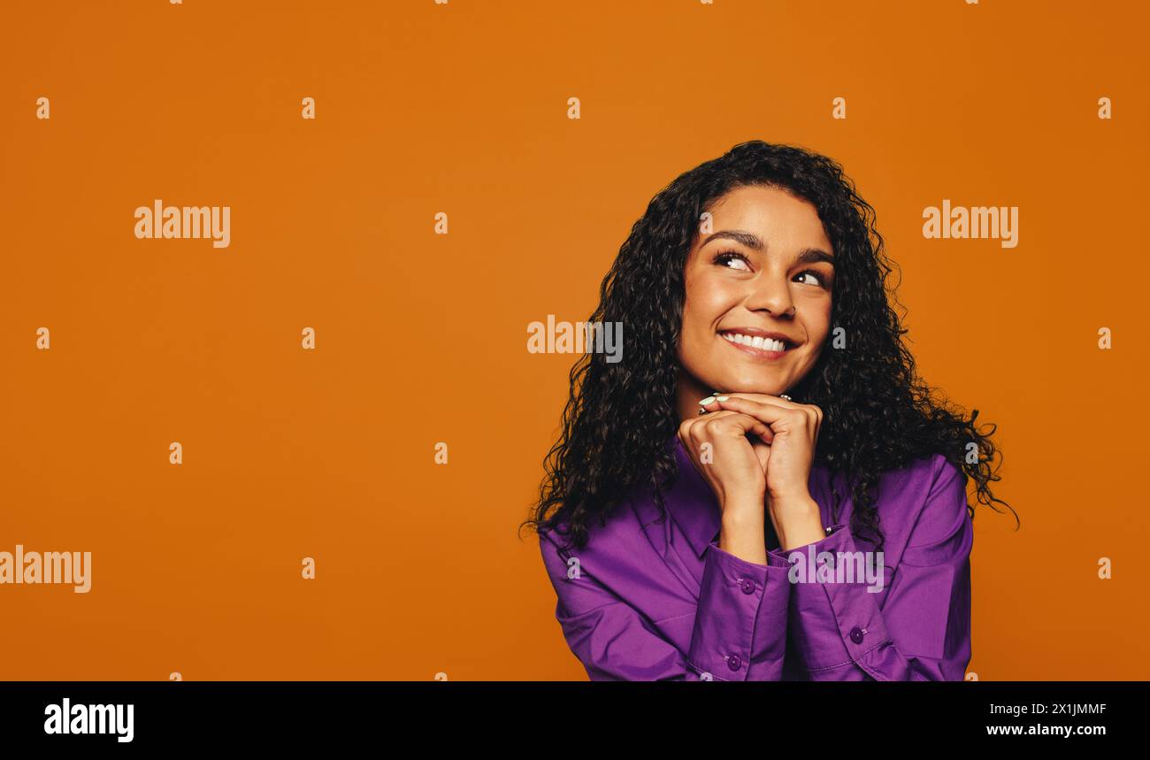 Auf einem orangefarbenen Hintergrund blickt eine fröhliche Frau mit lockigen Haaren und einem lebhaften Lächeln nachdenklich. Ihre farbenfrohe Kleidung trägt zu ihrem Jugendlichen und lässigen V Stockfoto