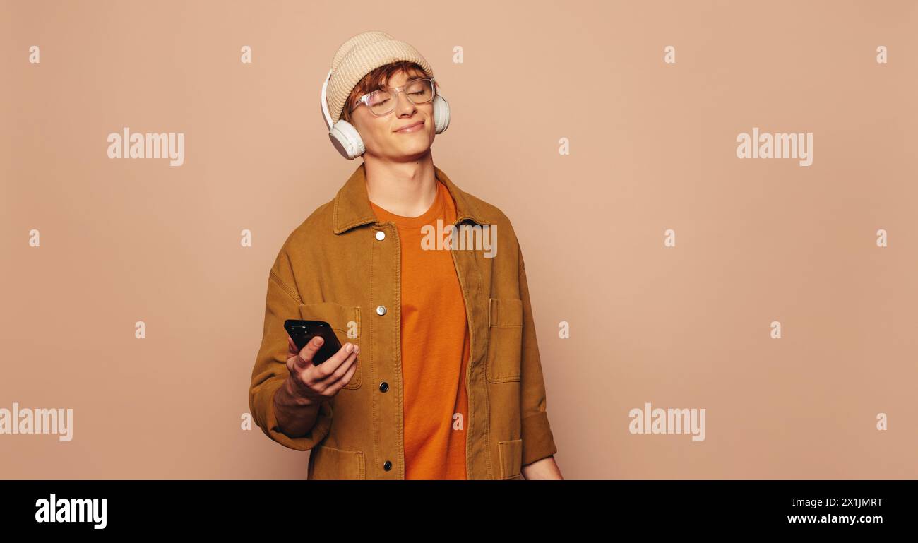 Der junge Mann hält ein Smartphone und trägt eine Brille, eine Jeansjacke und lässige Kleidung vor einem lebhaften pfirsichfarbenen Hintergrund, um Musik zu hören Stockfoto