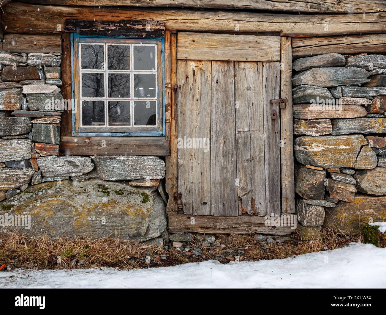 Tür und Fenster aus verwittertem Holz in Steinfundament inmitten von Schneespuren. Stockfoto