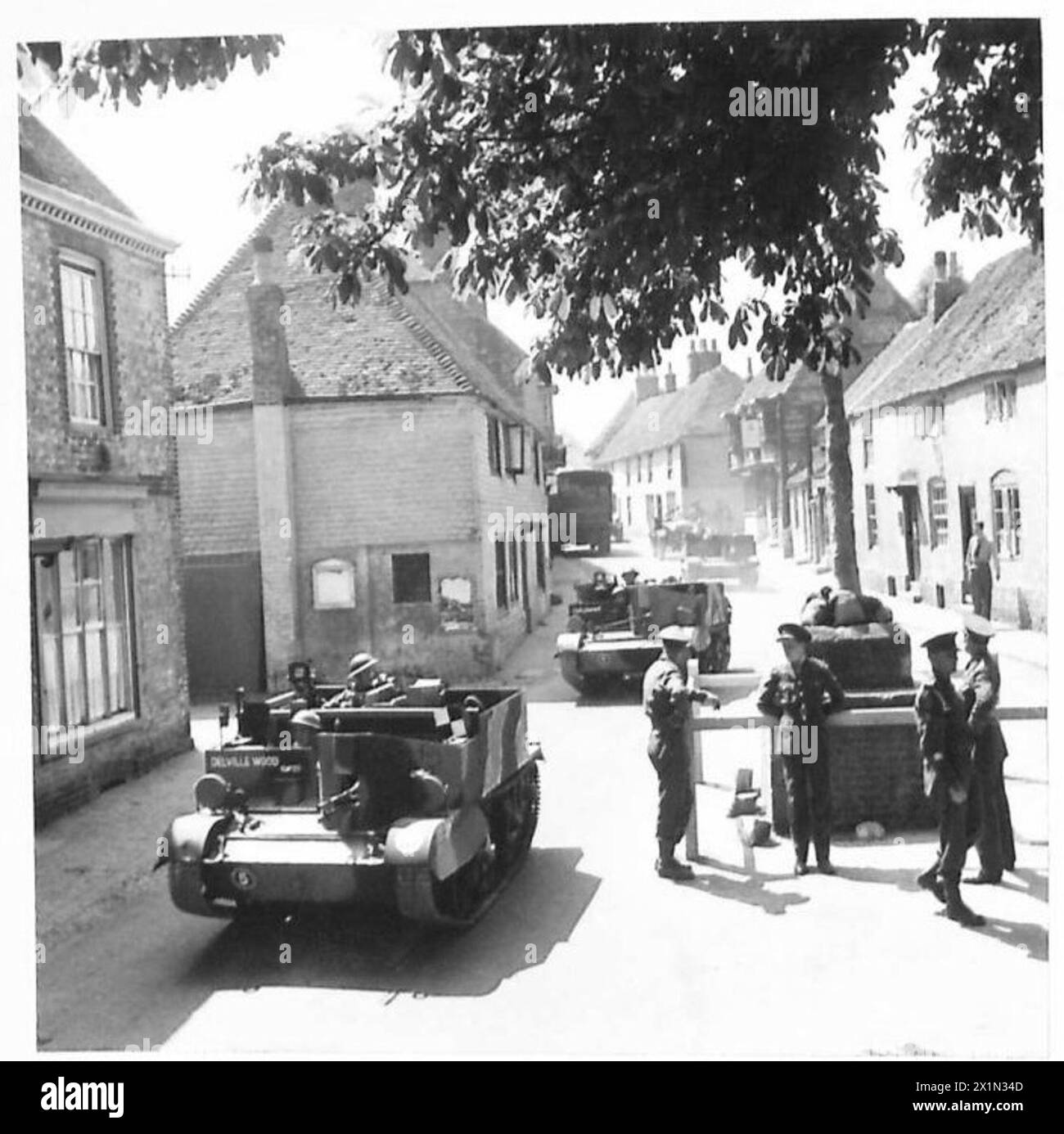 Bren-Träger der britischen Armee fahren auf dem Weg zum Bahnhof durch ein Dorf, um moderne Fahrzeuge mit einem alten Marktkreuz zu verbinden. Stockfoto