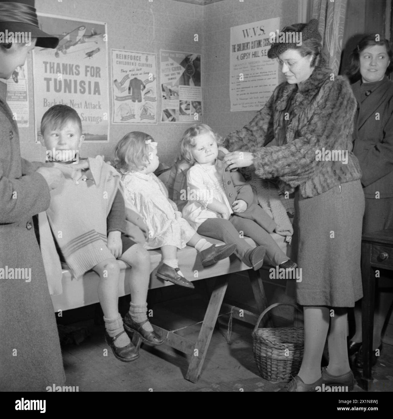 Bei einer WVS-Kinderkleidung-Börse in Norwood, London, im Jahr 1943 hilft Mrs. Gosmay ihrer Tochter Celia, einen Mantel anzuprobieren, während andere Kinder und Familienmitglieder teilnehmen. Stockfoto