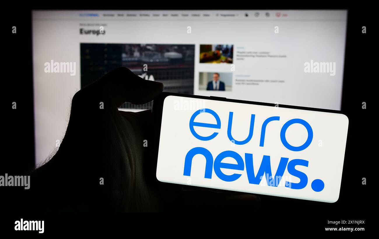 Person, die ein Mobiltelefon mit dem Logo des belgischen Nachrichtenunternehmens Euronews SA vor der Website des Unternehmens hält. Schwerpunkt auf dem Display des Telefons. Stockfoto