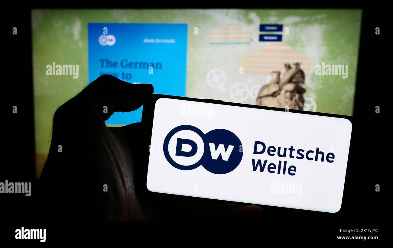 Person mit Mobiltelefon mit Logo der Deutschen Welle (DW) vor der Webseite. Schwerpunkt auf dem Display des Telefons. Stockfoto