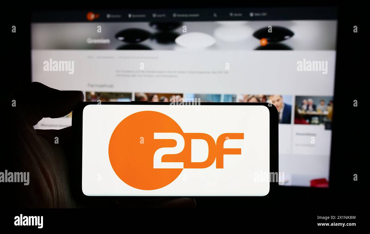 Person, die ein Mobiltelefon mit dem Logo des Fernsehsenders zweites Deutsches Fernsehen (ZDF) vor der Webseite hält. Schwerpunkt auf dem Display des Telefons. Stockfoto