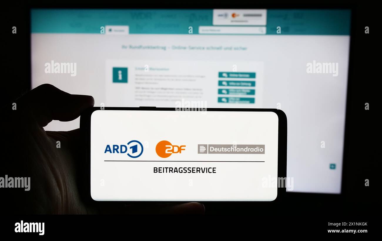 Person mit Handy mit Logo des ARD ZDF Deutschlandradio Beitragsservice vor der Webseite. Schwerpunkt auf dem Display des Telefons. Stockfoto