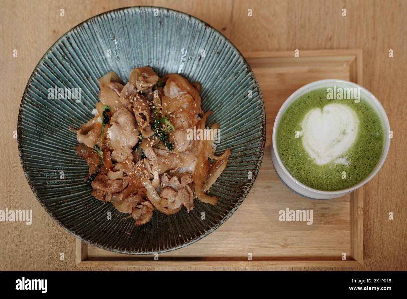 Schweinebraten mit Gemüse, bestreut mit Sesamsamen, gepaart mit einer cremigen Matcha-Grüntee-Latte, kunstvoll mit einem herzförmigen Schaum garniert, Stockfoto