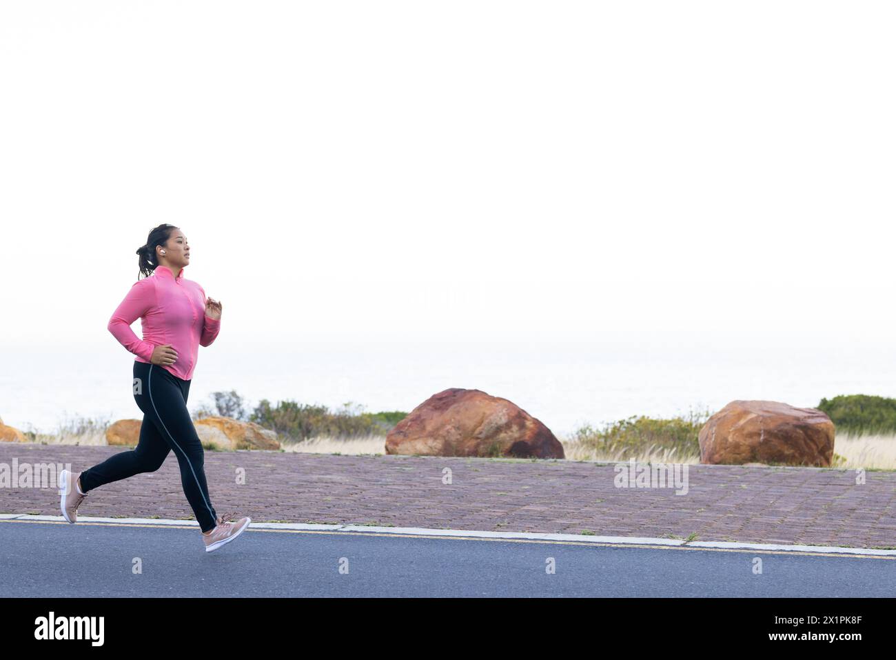 Eine birassische Wanderin joggt am Meer in rosa Oberteil und Leggings, Kopierraum Stockfoto