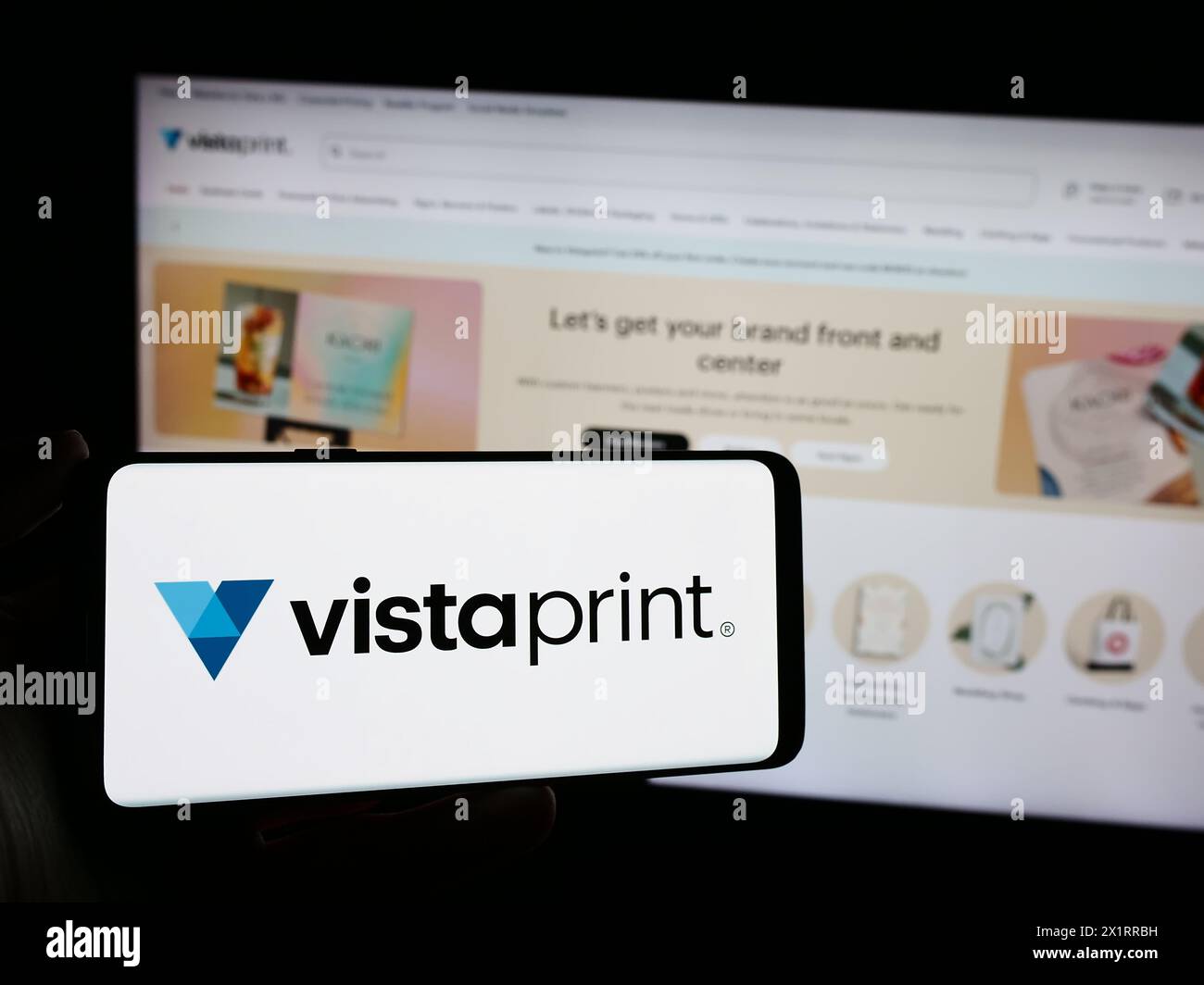 Person, die ein Mobiltelefon mit dem Logo des Unternehmens Vistaprint (Vista) vor der Unternehmenswebseite hält. Schwerpunkt auf dem Display des Telefons. Stockfoto