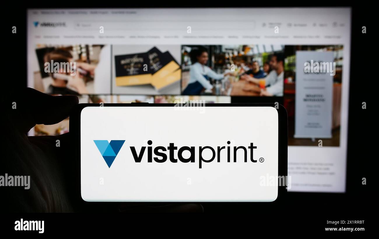 Person, die ein Mobiltelefon mit dem Logo des Unternehmens Vistaprint (Vista) vor der Unternehmenswebseite hält. Schwerpunkt auf dem Display des Telefons. Stockfoto