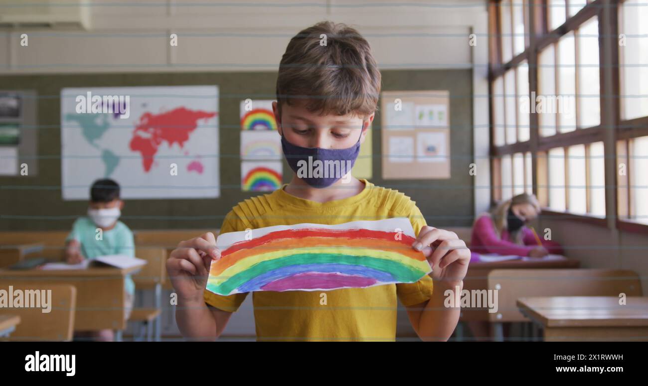 Ein kaukasisches Kind in gelbem Hemd und Maske zeigt in der Schule eine Regenbogenzeichnung Stockfoto