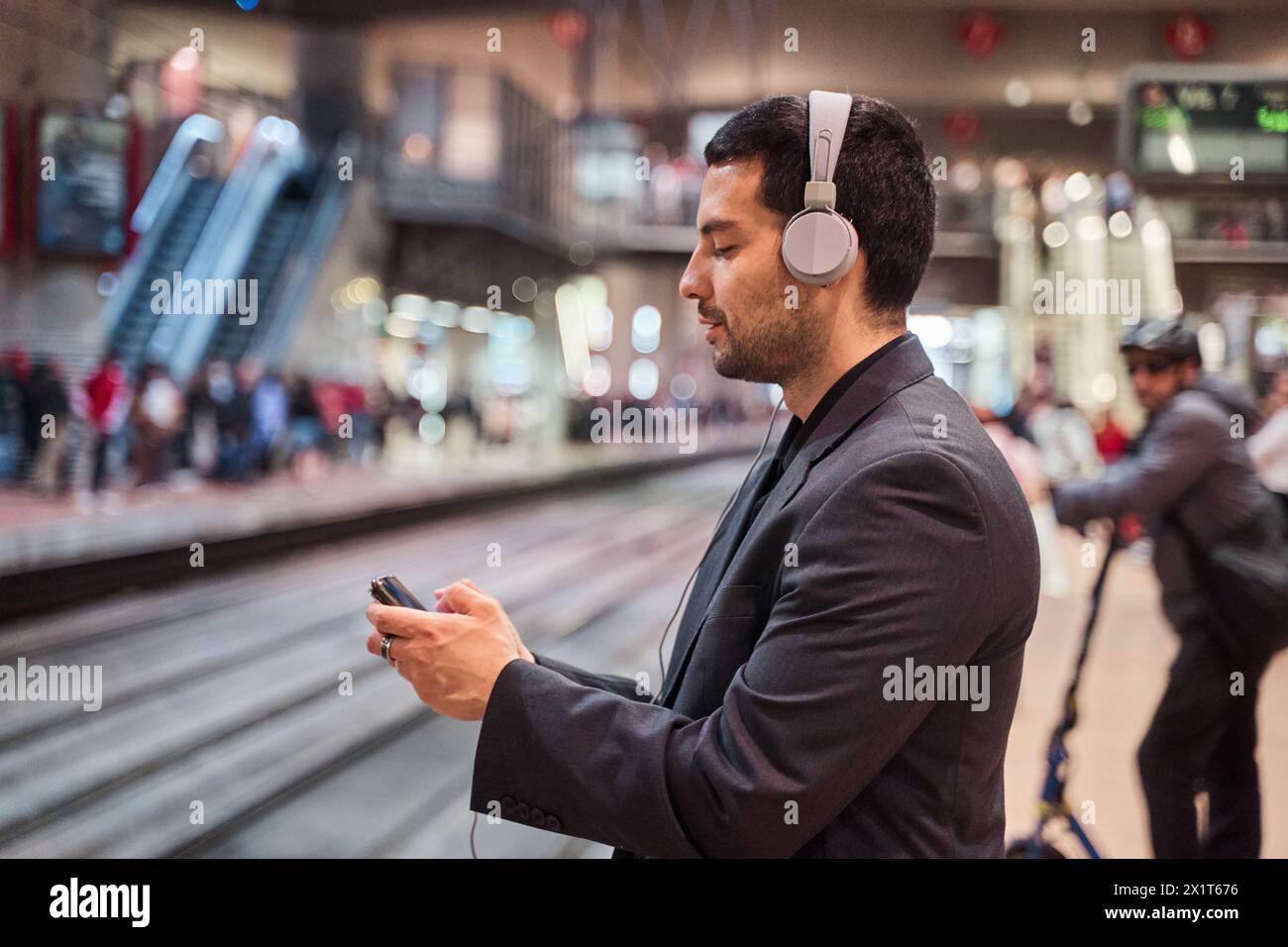 latein-Mann, der auf den Zug wartet, während er sein Smartphone mit Kopfhörern benutzt, um Spiele zu spielen oder ein Video anzusehen Stockfoto