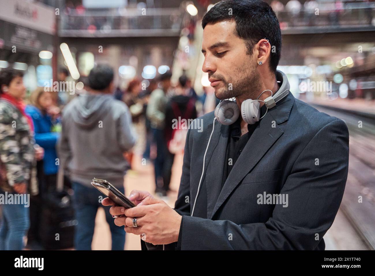 latino-Geschäftsmann wartet am Bahnhof und schaut auf das Smartphone mit Leuten im Hintergrund Stockfoto