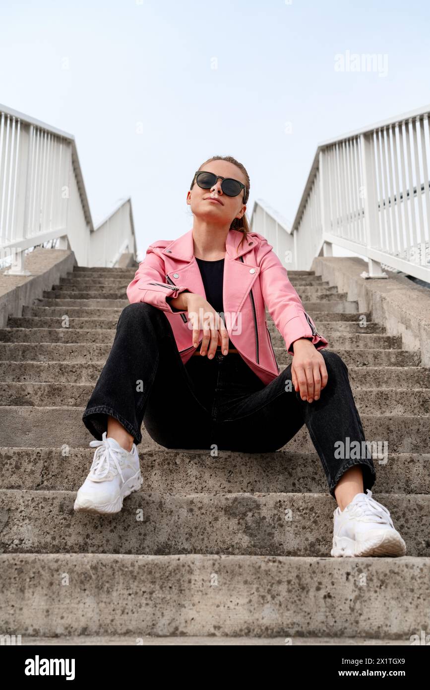 Die modische, stilvolle Frau in einer rosa Kunstlederjacke und Sonnenbrille sitzt auf den Stufen einer Stadtstraße. Stockfoto