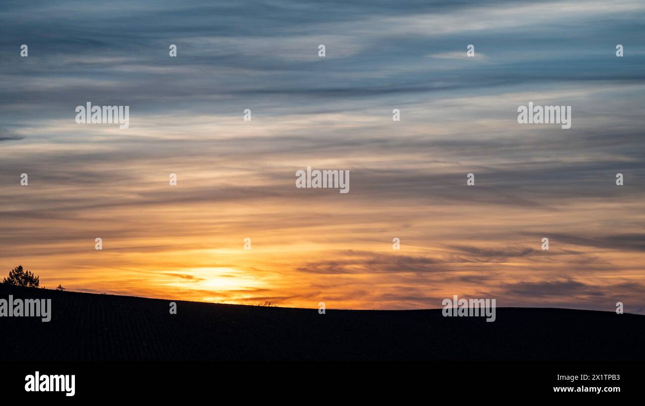 Sonnenuntergang über einer friedlichen Landstraße, mit dem Himmel in warmen Orange- und Gelbtönen gemalt, führt zu einem ruhigen Bauernhaus in der Ferne. Stockfoto