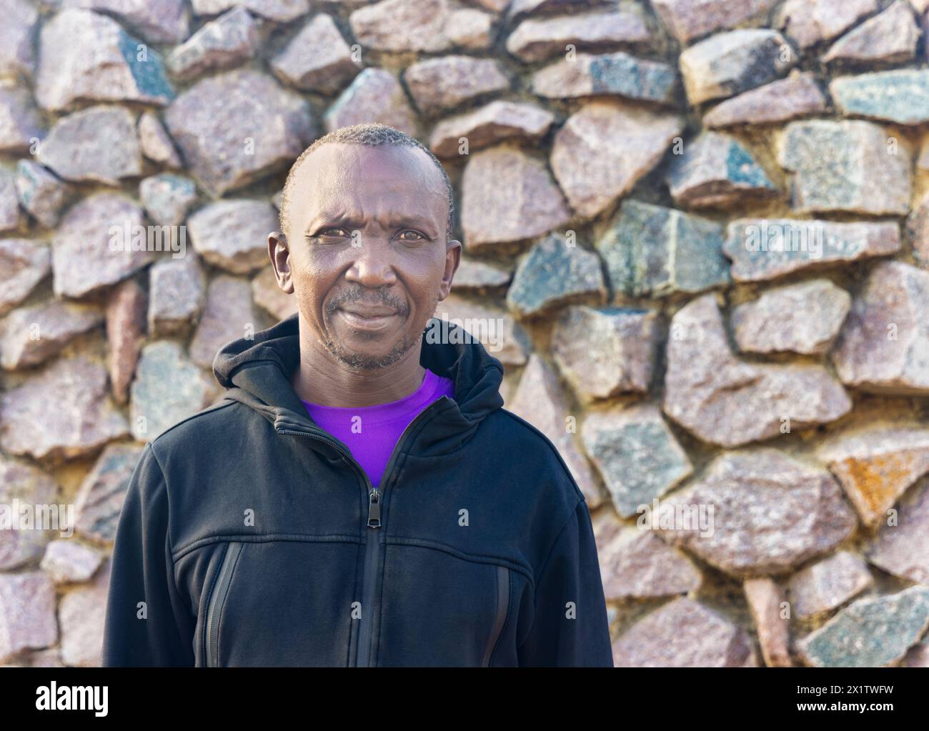 afrikanischer Mann mit mittlerem Erwachsener, der mit einem schwarzen Hoodie gekleidet ist, draußen an einem sonnigen Tag vor einer Steinmauer Stockfoto