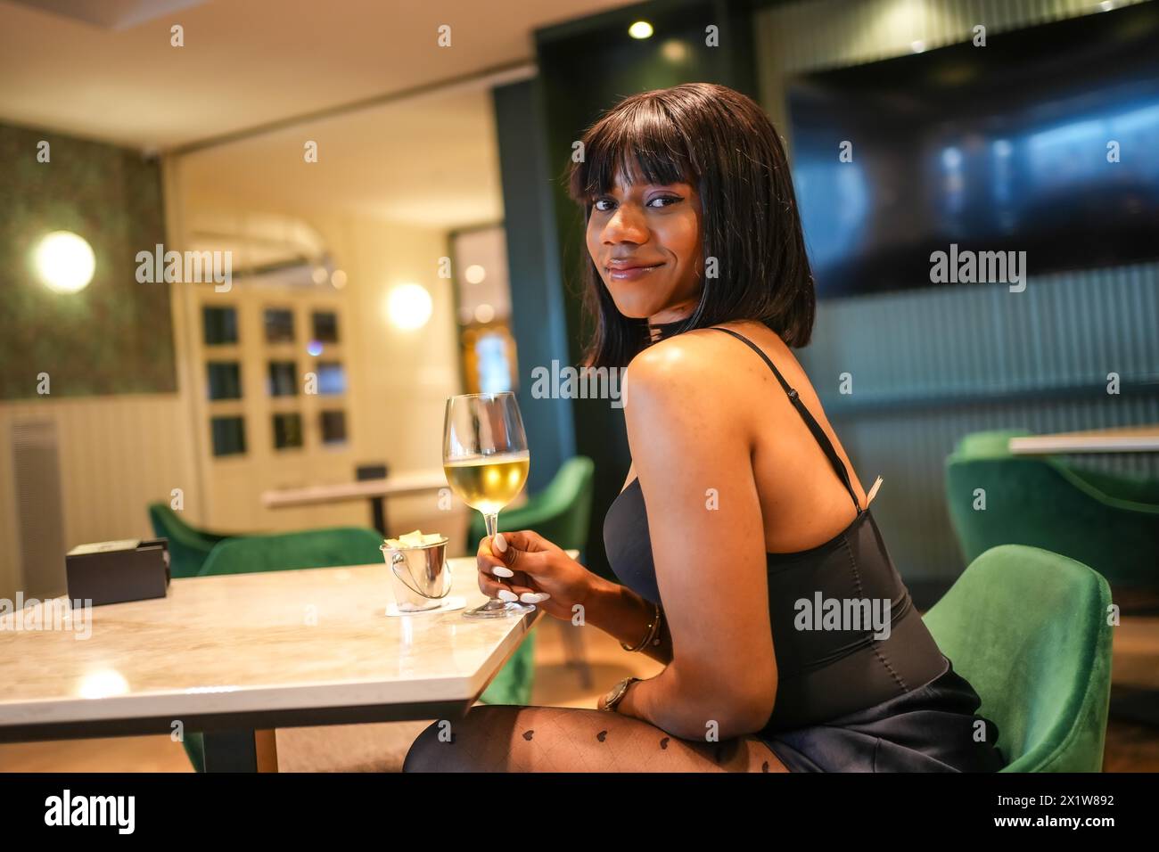 Afrikanische junge Frau lächelt in die Kamera, während sie in einer glamourösen Bar Weißwein trinkt Stockfoto