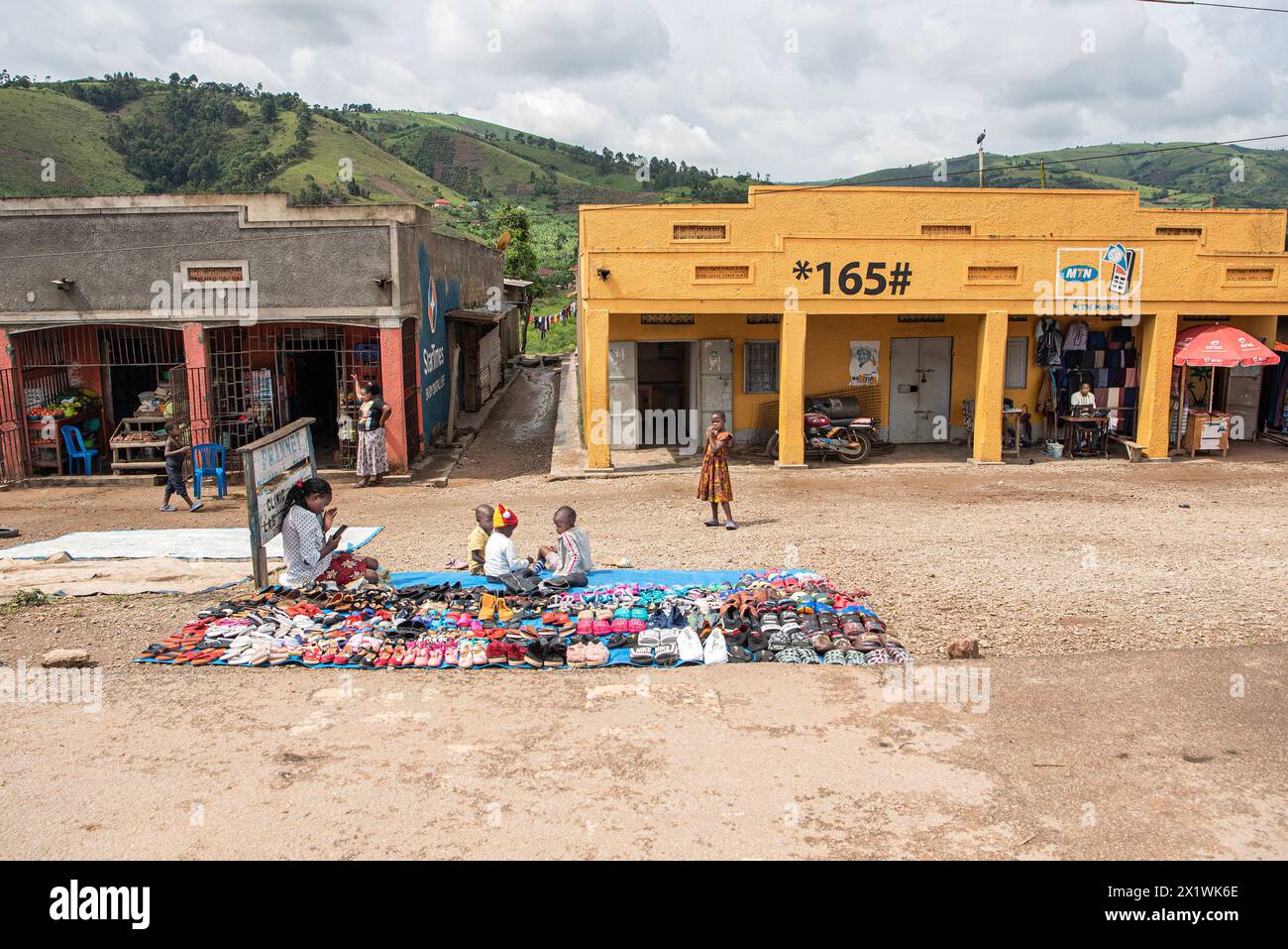 In einem energiegeladenen ugandischen Dorf drängen Händler die Straßen mit farbenfrohen Displays. Kinder tummeln sich in der Nähe als Frau, umgeben von einer Vielzahl von Snea Stockfoto