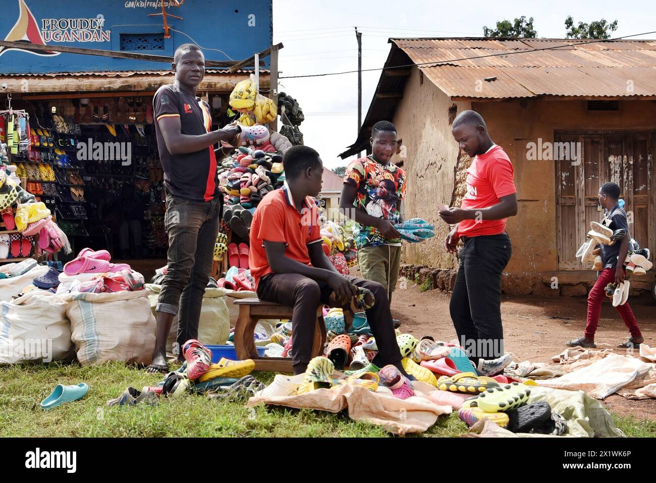 Eine einfache Lehmhütte steht neben einem bescheidenen ugandischen Schuhladen am Straßenrand. Drei Jugendliche verkaufen Turnschuhe aus großen weißen Säcken in der Nähe. Stockfoto